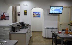 A compact kitchen with a stove, microwave, and cabinets, leading to a dining area with a table and chairs. A TV is mounted on the wall displaying 'LA CHIOCOLA APARTMENT'.