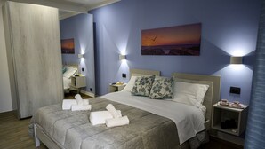 A neatly made bed with a grey quilt, two pillows, and rolled towels on a wooden floor. There are two bedside tables with lamps, a wall-mounted mirror, and a painting of a sunset.