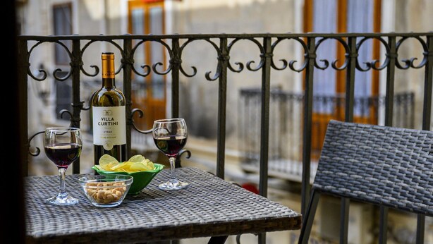 A balcony with a table set for two, featuring a bottle of Villa Curtini wine, two glasses, a bowl of chips, and a bowl of nuts.