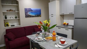 A compact living space with a dining area, a sofa, a kitchen with a refrigerator, and a wall-mounted shelf with decorative items.