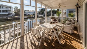 Een overdekt terras buiten met houten vloer, witte meubels en uitzicht op een jachthaven met boten.