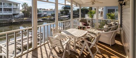 Een overdekt terras buiten met houten vloer, witte meubels en uitzicht op een jachthaven met boten.