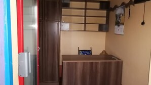 A room with a wooden wardrobe, a desk with a chair, and a wall-mounted shelf.