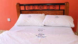 A bed with white bedding and pillows, a wooden headboard with a decorative metal design, and a wall with a power outlet.