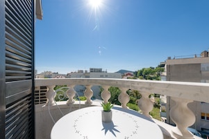 Un balcon agrémenté d’un pot de fleurs, d’une table blanche et offrant une vue dégagée sur les bâtiments et le paysage environnants.