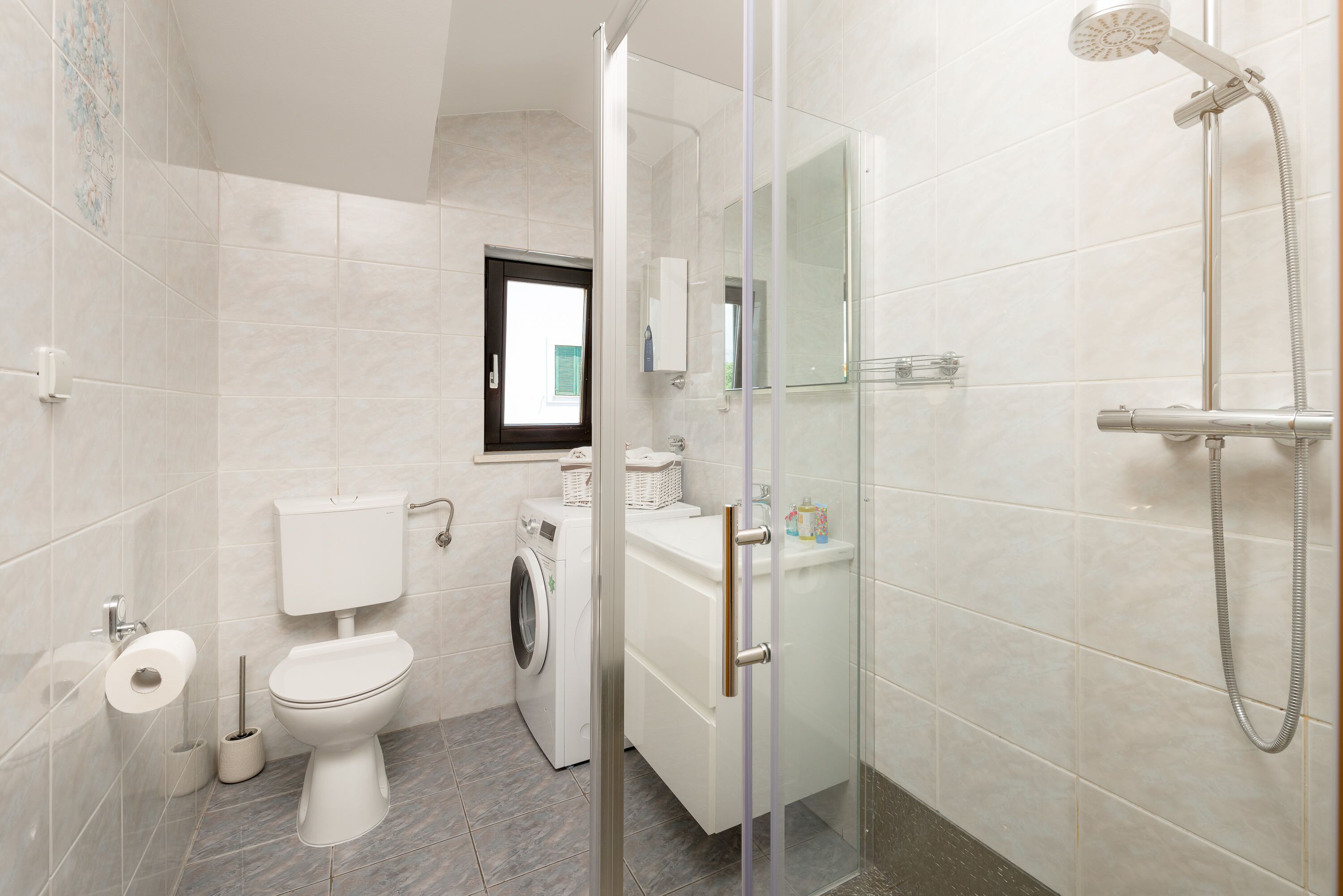 A modern bathroom with a toilet, washing machine, and glass-enclosed shower.