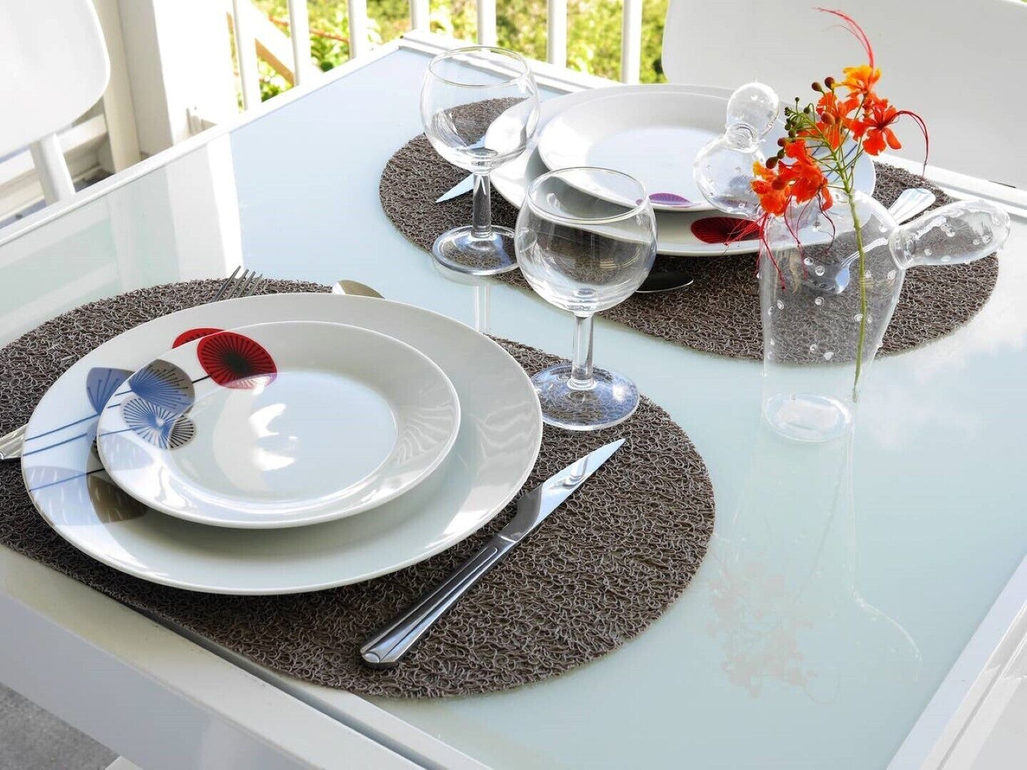 Mesa posta com pratos, talheres e copos de vinho sobre uma mesa de vidro, com vista para o verde lá fora.