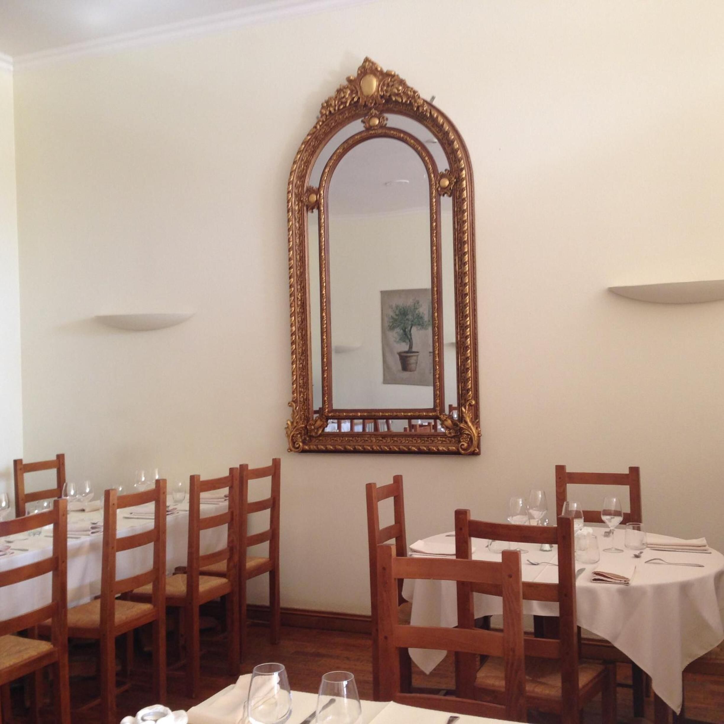 Un comedor con sillas y mesas de madera, un espejo grande y ornamentado en la pared, y una planta en maceta en el reflejo.
