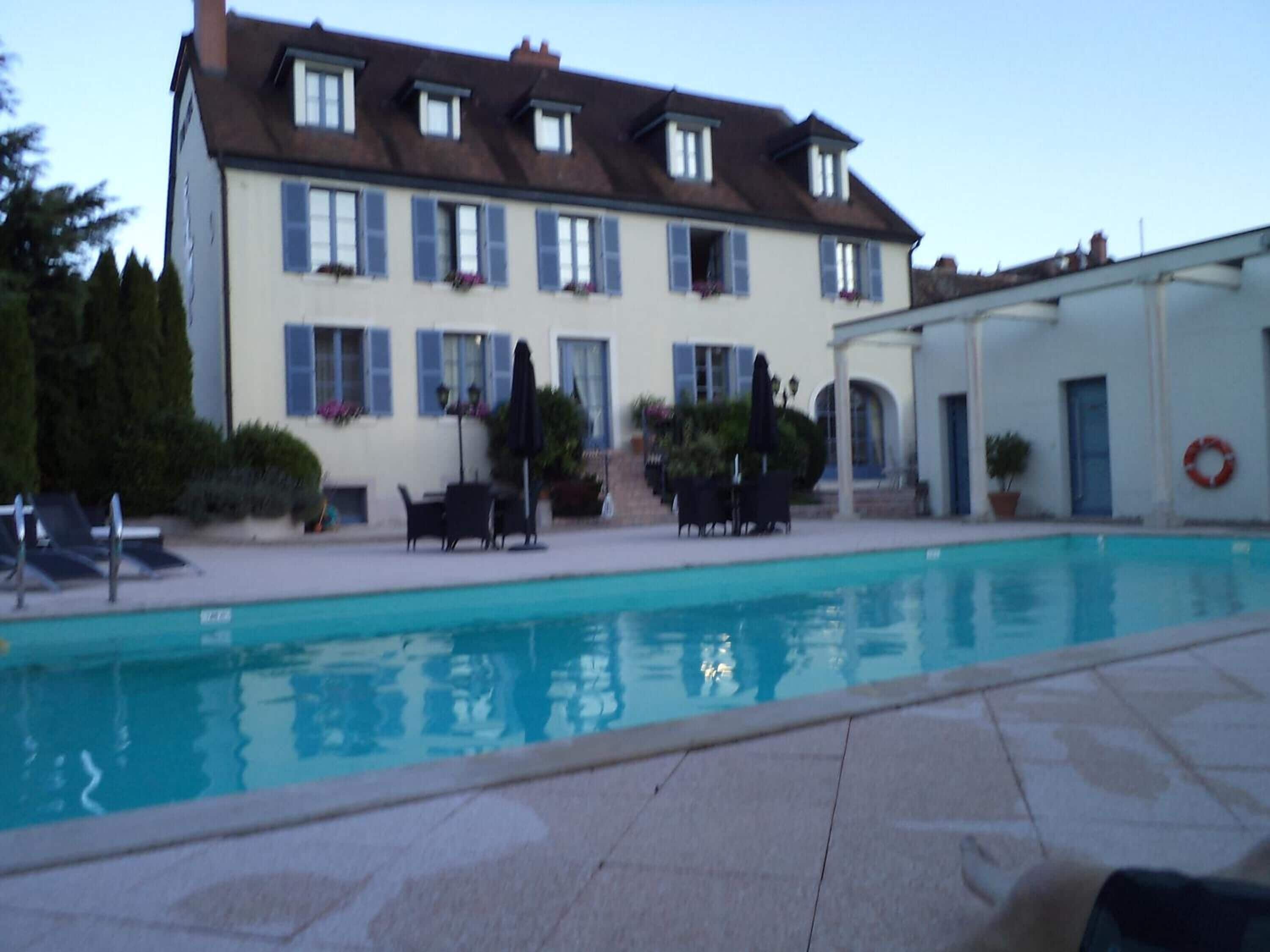 A swimming pool with lounge chairs and umbrellas, a building with multiple windows and shutters, and a lifebuoy on the wall.
