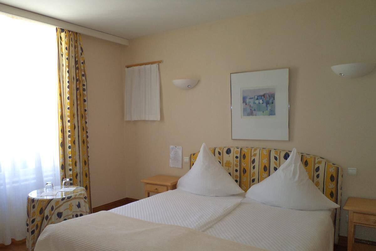 A hotel room with a bed, a bedside table, a small round table, a window with curtains, and a wall-mounted picture.