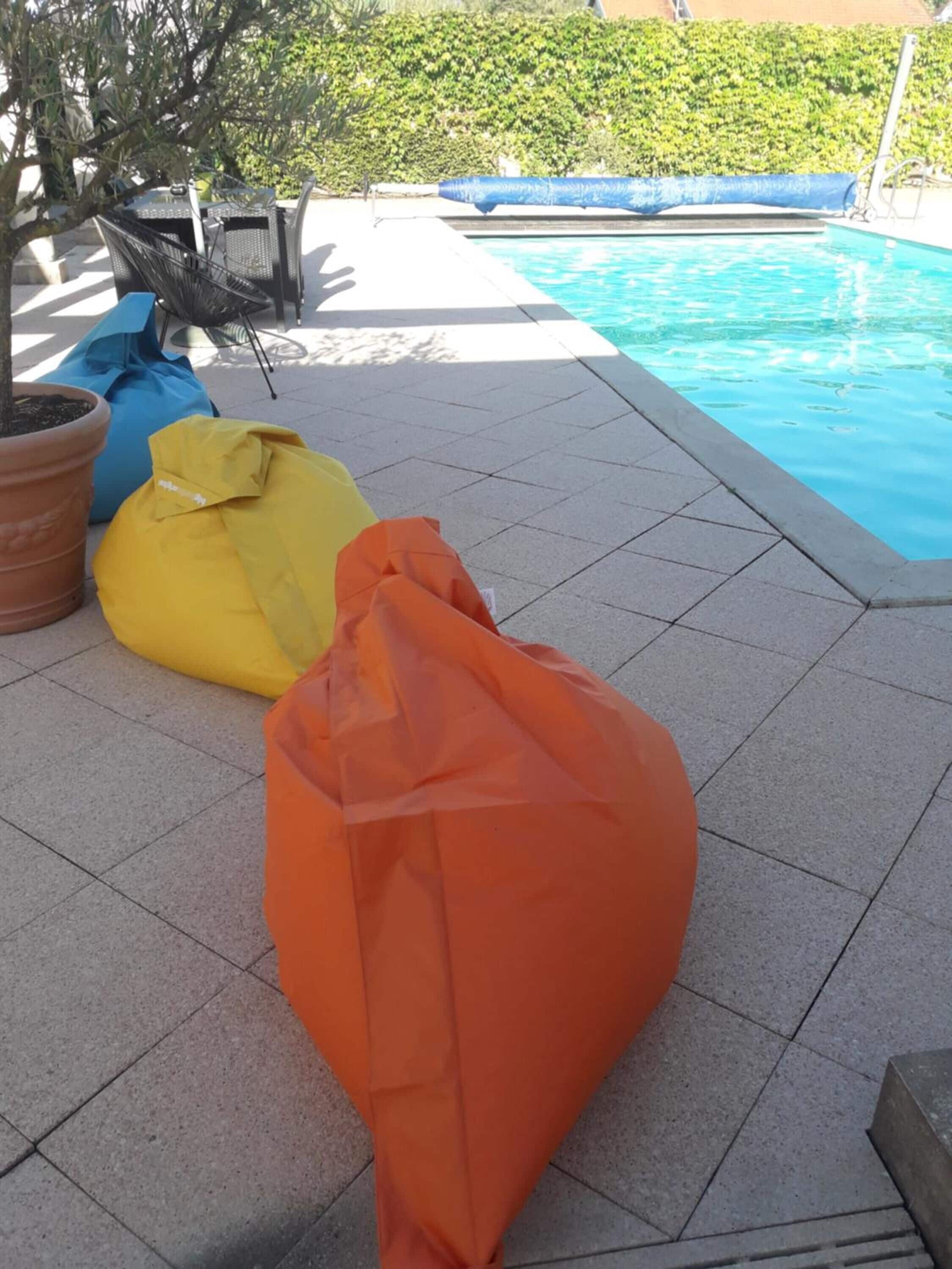 A poolside area with a tiled patio, a blue pool, and three bean bags in orange, yellow, and blue colors.