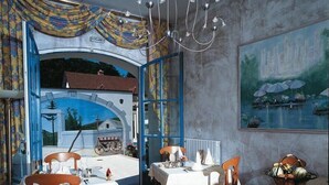 A dining area with tables set for two, a chandelier, and a view of a rural landscape through an open archway.