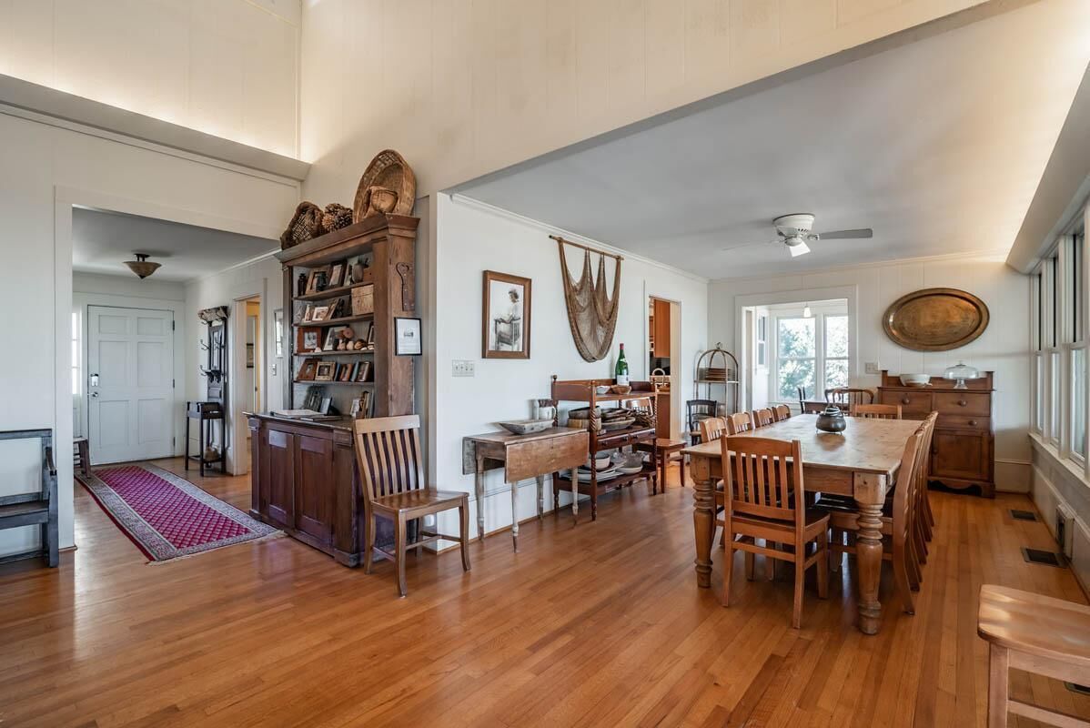 A spacious room with wooden flooring, a dining table with chairs, a bookshelf, and a large window.