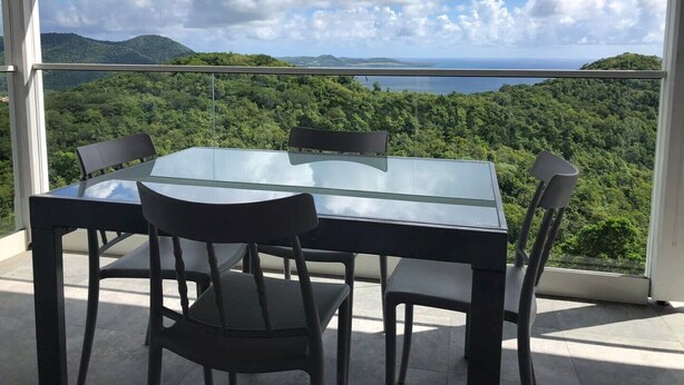 Ein Tisch mit Glasplatte und vier Stühlen auf einem Balkon mit Blick auf eine üppige grüne Landschaft, Berge und das Meer im Hintergrund.