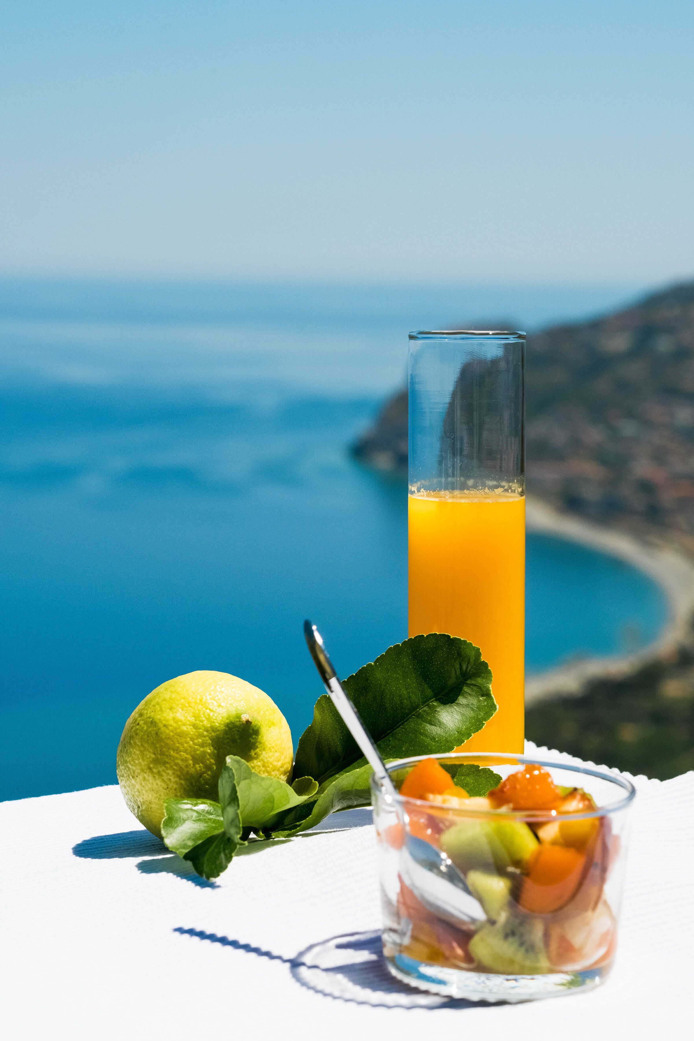 A glass of orange juice with a lemon and mint leaves on a table with a scenic coastal view in the background.