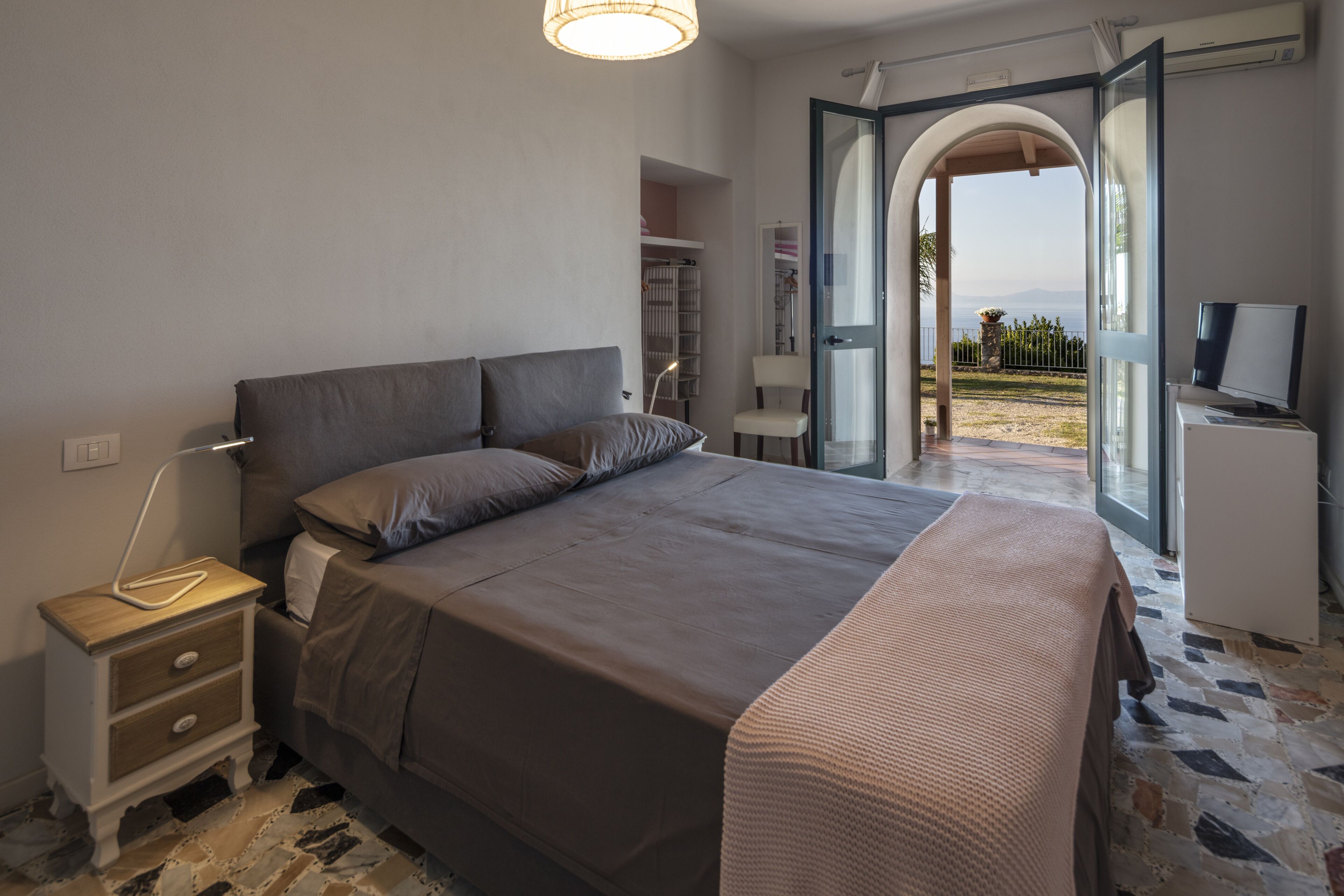 Une chambre à coucher avec un lit, une table de chevet, une lampe et une porte ouverte donnant sur un balcon.