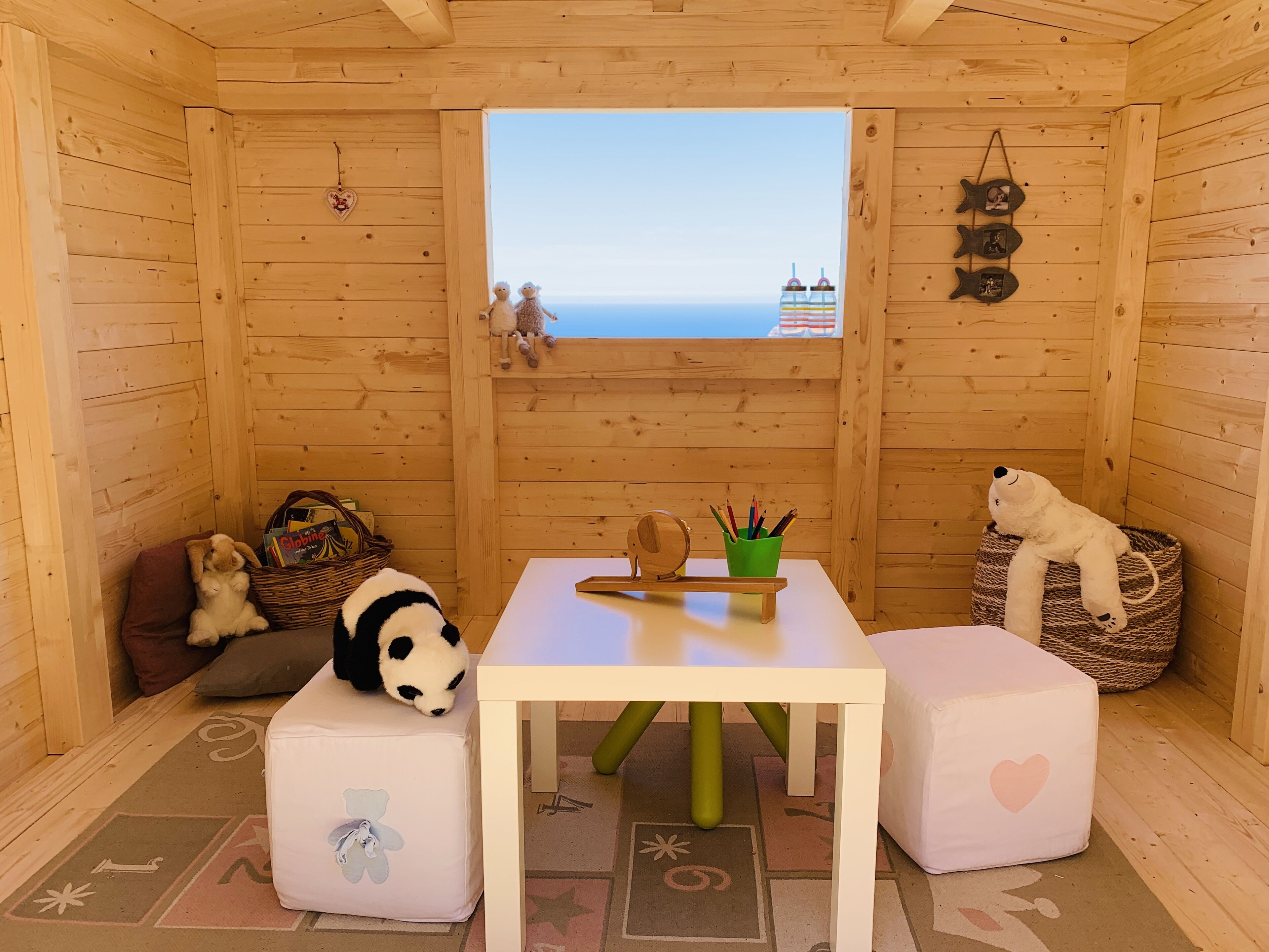 A cozy wooden room with a view of the sea, featuring a table with a green cup holder, a panda plush toy, and a polar bear plush toy.