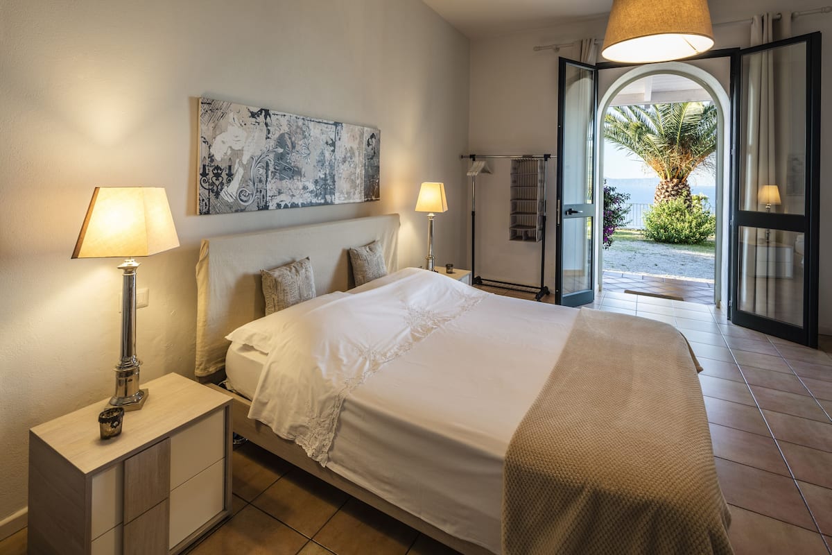 Een slaapkamer met een groot bed, twee bedlampen, een nachtkastje, een aan de muur hangend kunstwerk en een open deur naar een balkon met uitzicht op zee.