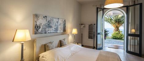 A bedroom with a large bed, two bedside lamps, a nightstand, a wall-mounted artwork, and an open door leading to a balcony with a view of the sea.