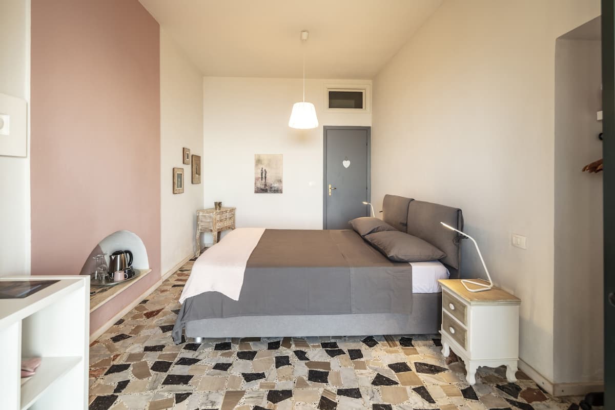 Een slaapkamer met een bed, nachtkastje, lamp en een wandplank met waterkoker en kopjes.