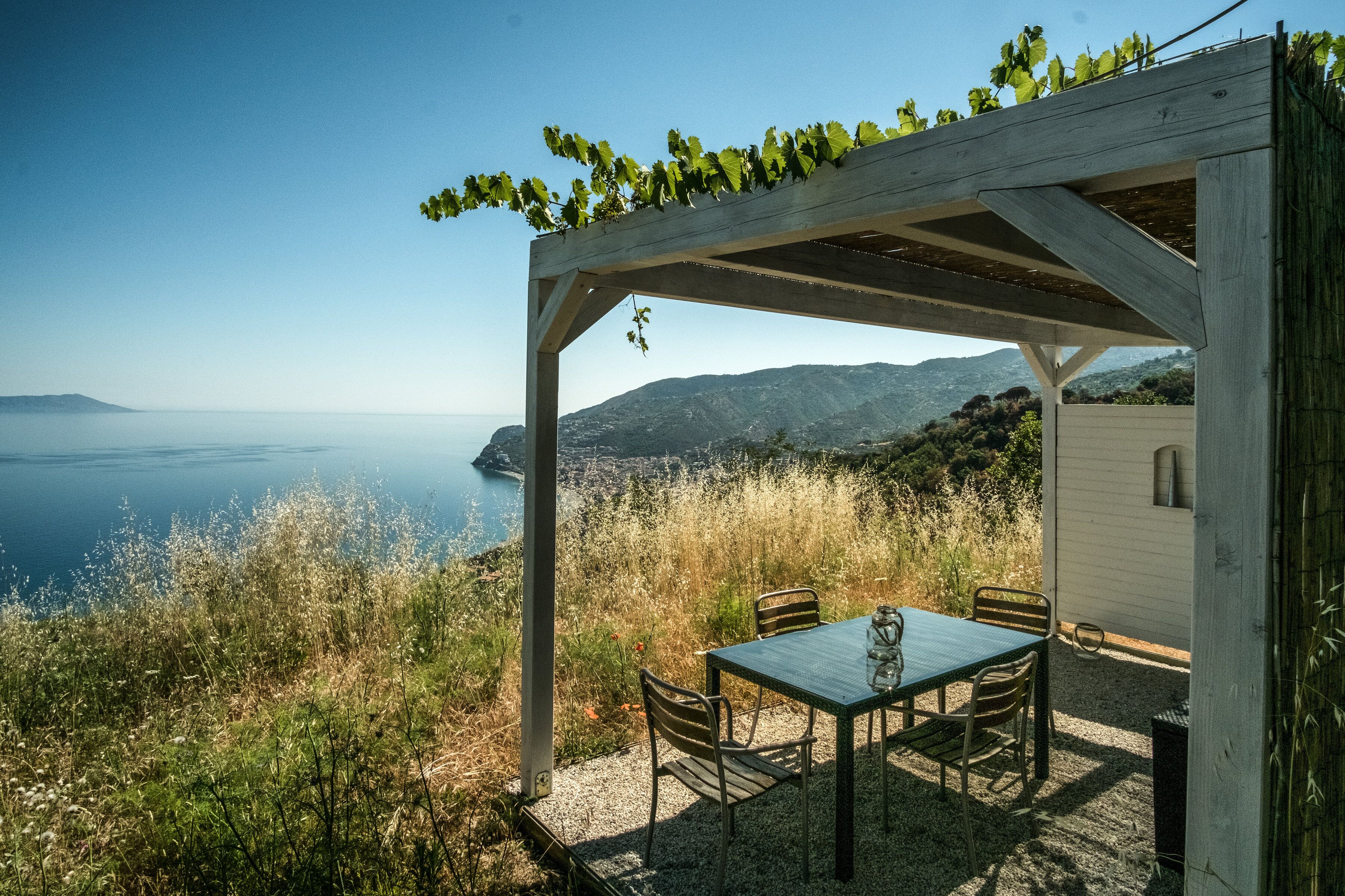 A wooden pergola with a table and chairs overlooking a coastal landscape.