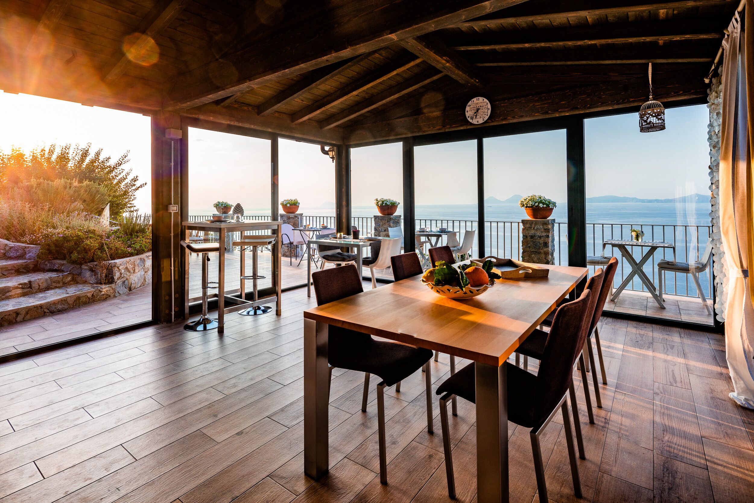 A spacious dining area with wooden flooring and tables, large windows offering a view of the sea and mountains, and potted plants on the tables.