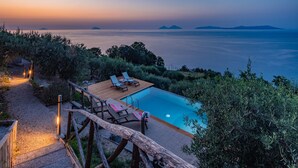 Une terrasse en bois avec deux chaises longues et une petite table, donnant sur une piscine et la mer.