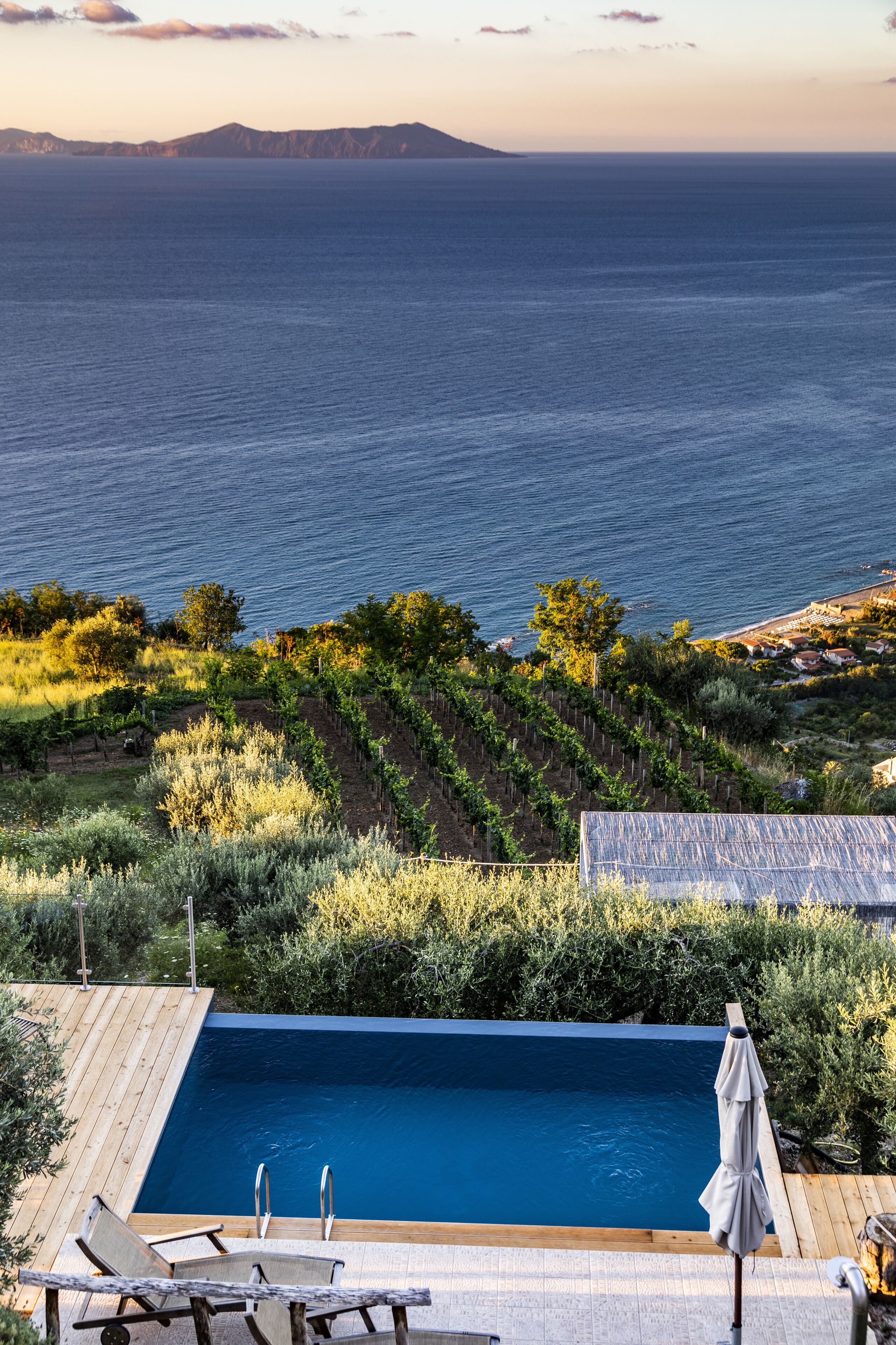 Piscina con muelle de madera y sillones de descanso, rodeada de vegetación y con vista a una masa de agua y formas de relieve en la distancia.