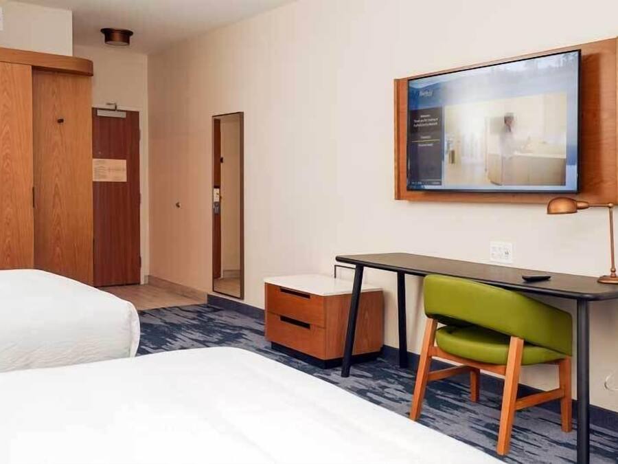 A hotel room with two beds, a desk with a chair, a small table, a mirror, and a television mounted on the wall.