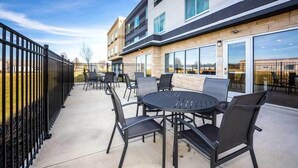 Un espace extérieur aménagé en terrasse, avec une table ronde et des chaises, une clôture en métal noir et un bâtiment doté de grandes fenêtres et d’une façade en pierre.