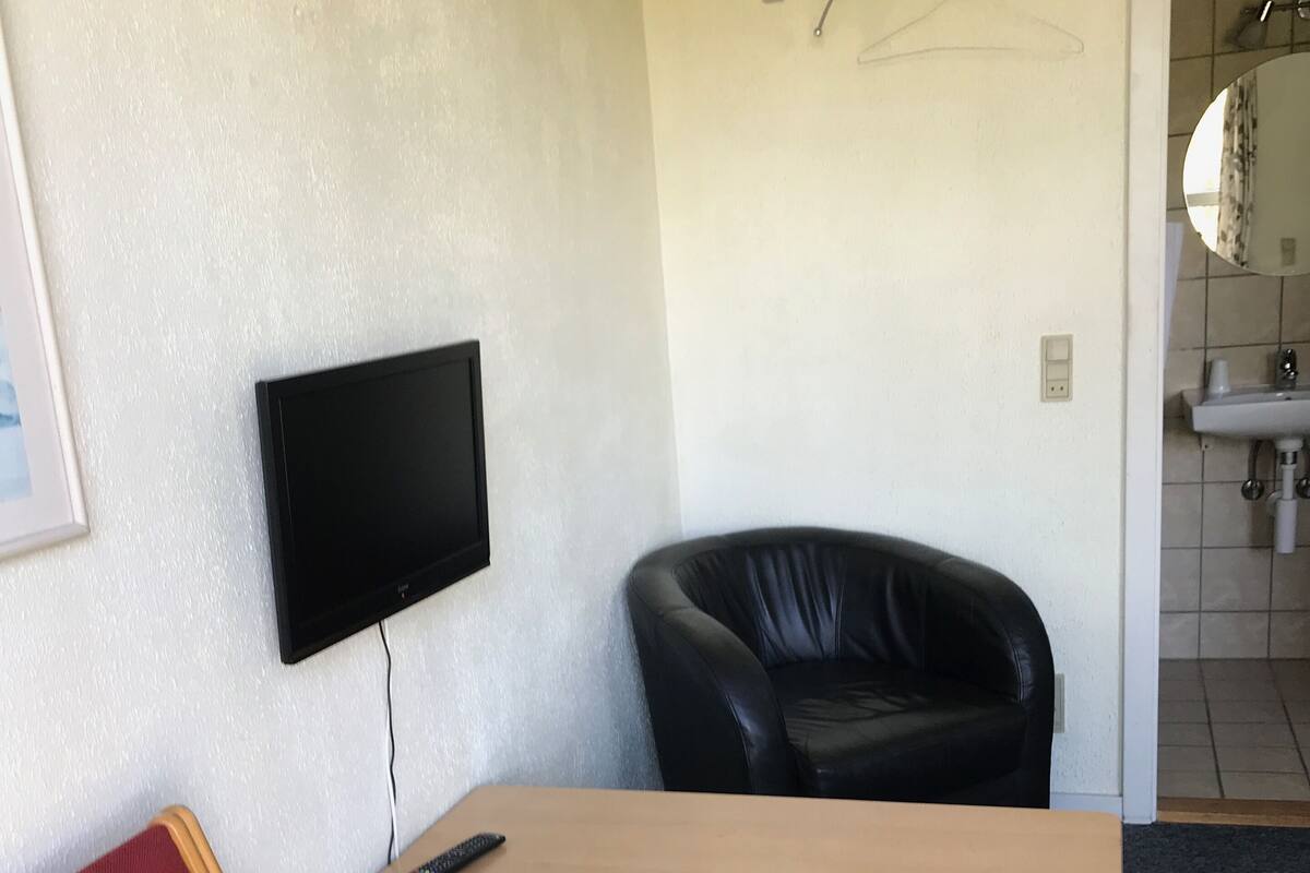 A room with a flat-screen TV mounted on the wall, a black leather chair, a wooden table, and a red chair.