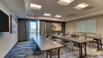 A conference room with long tables, chairs, a projector screen, and a large window with curtains.