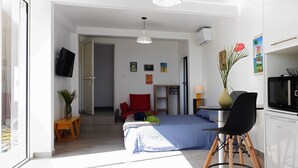 Une pièce compacte et bien éclairée, comprenant un lit, une petite table, une chaise et un coin cuisine.