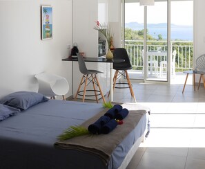 Une chambre à coucher avec un lit, un bureau et des chaises. Elle possède un balcon avec vue sur la mer.