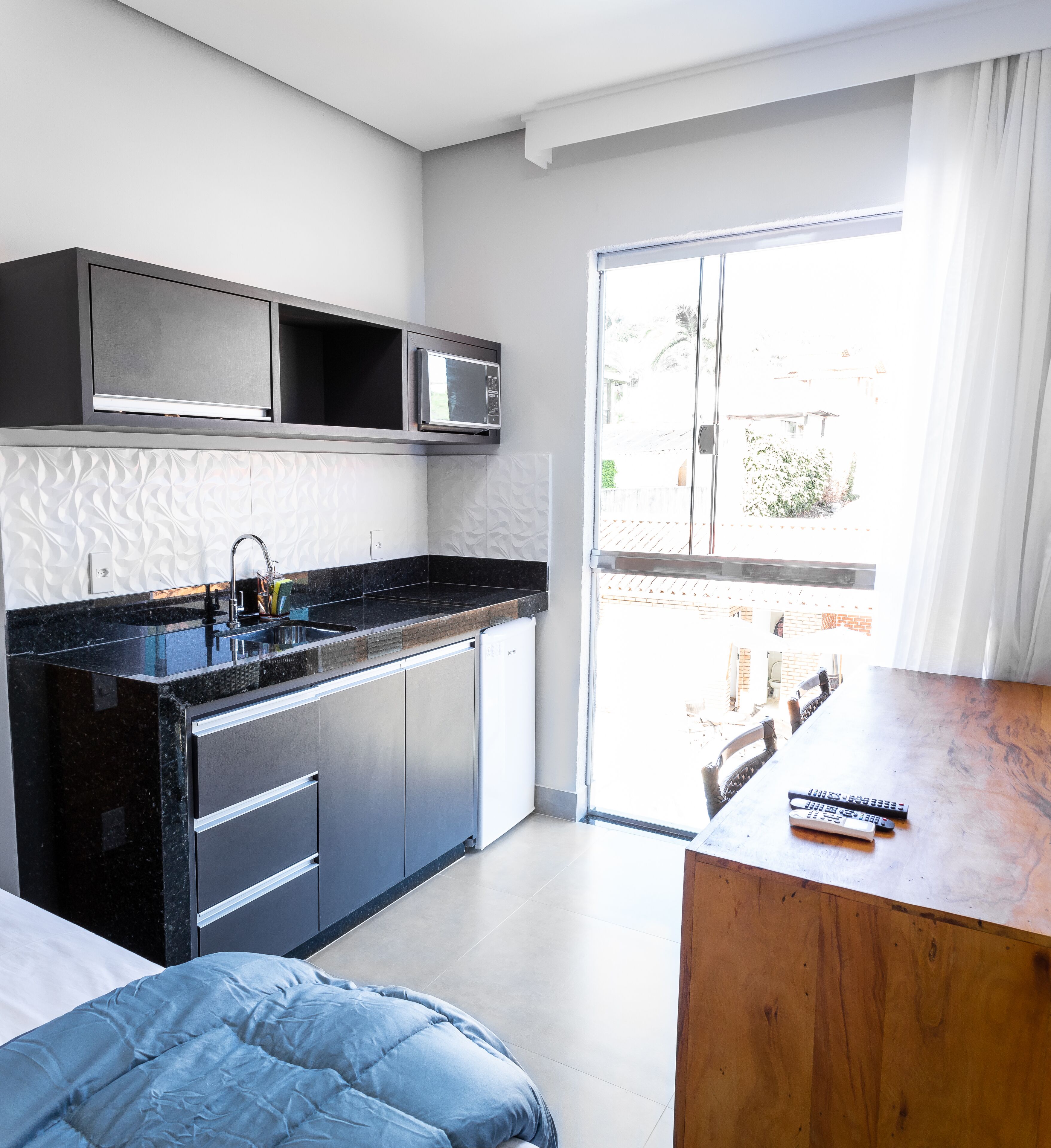 Een moderne keuken met een ingebouwde magnetron, een spoelbak en een vaatwasser. Er staat een houten tafel met afstandsbedieningen erop, en een bed met een blauwe deken staat op de voorgrond.