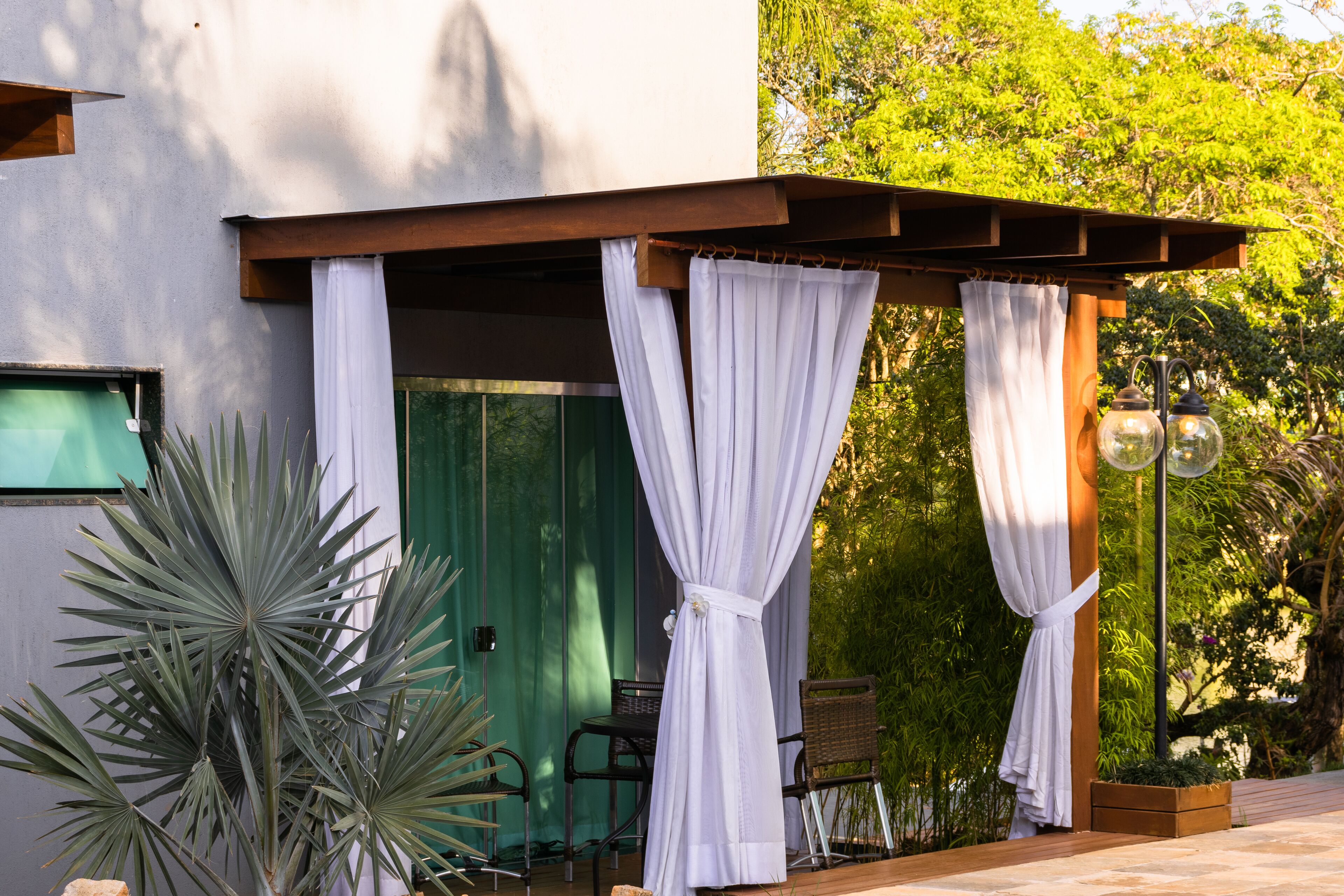 A modern outdoor seating area with a wooden pergola, white curtains, and a table with chairs.