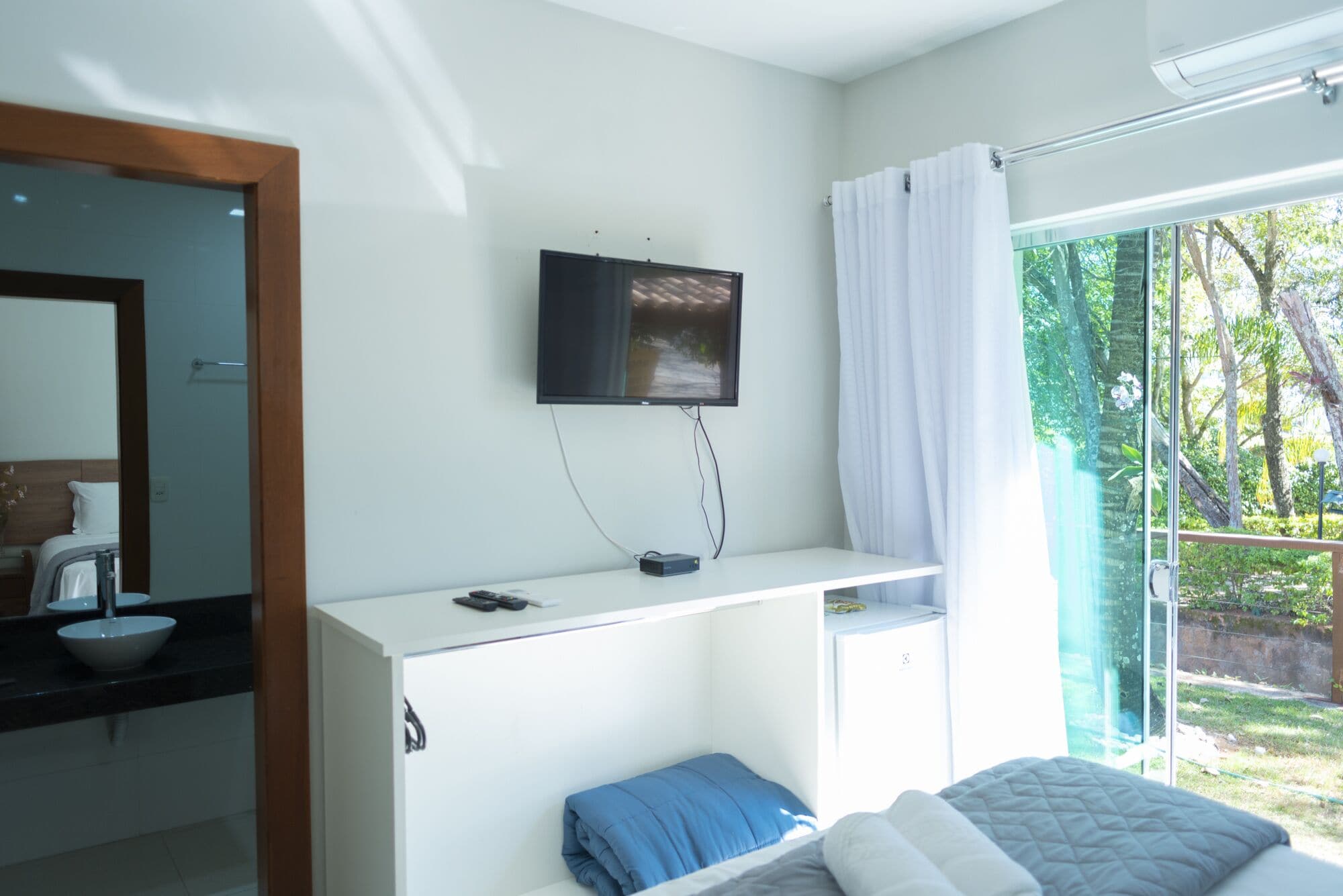 A modern hotel room with a flat-screen TV mounted on the wall, a small desk with a computer, a bed with a blue pillow, and a large window with a view of greenery.
