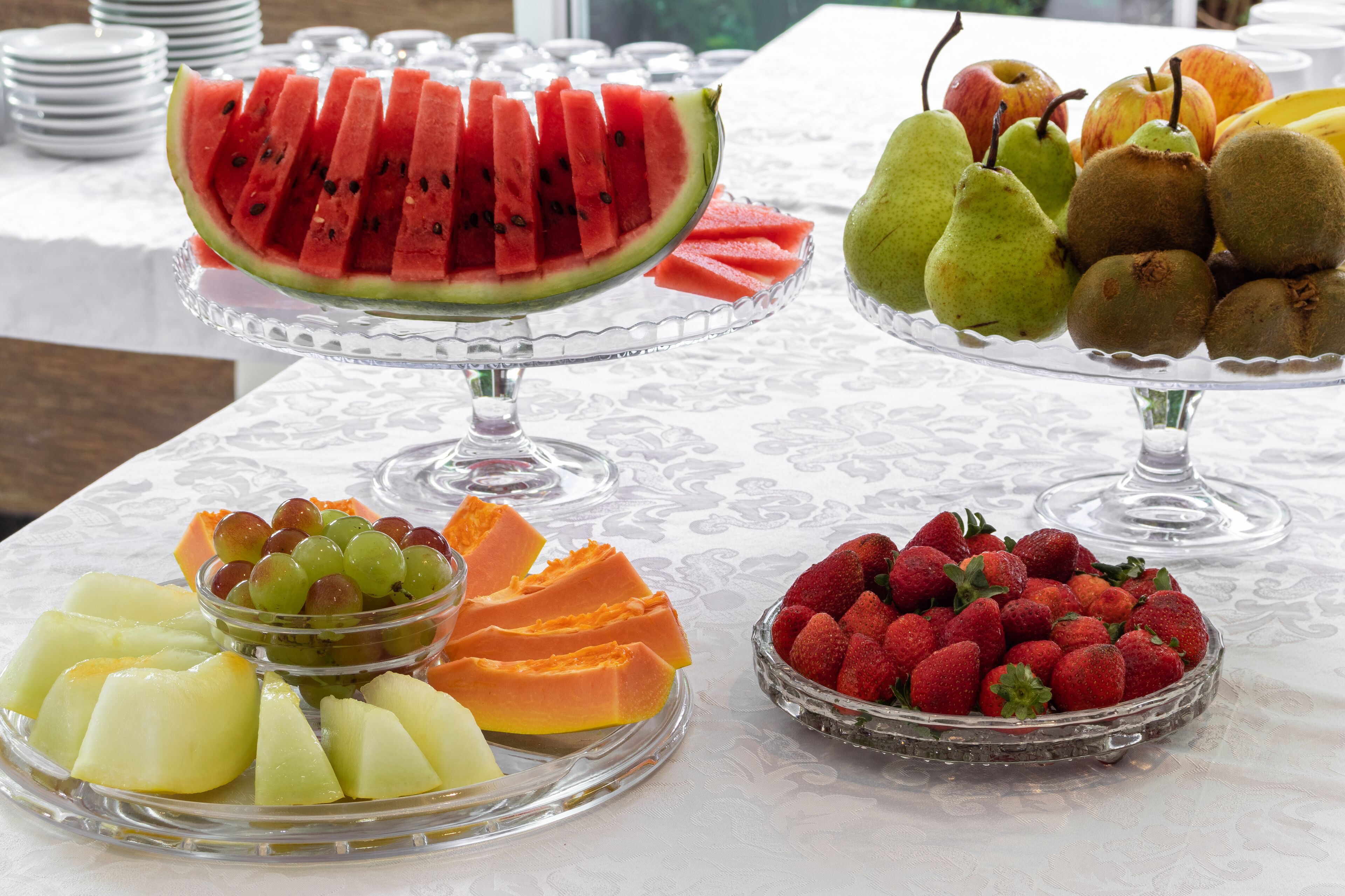 Een tafel met diverse fruitschaaltjes, waaronder watermeloen, peren, kippenfruit, druiven, papaja en aardbeien.