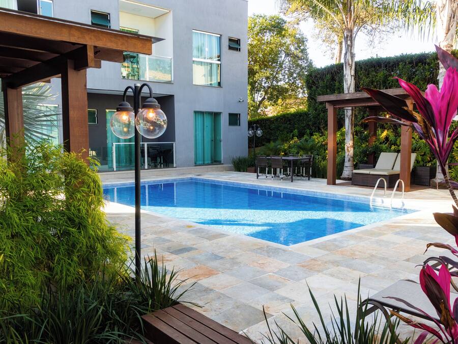 A swimming pool with a tiled deck, surrounded by greenery and a pergola.