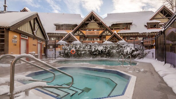 A pool area with a snow-covered deck, a lifeguard chair, and a building with a wooden facade and snow on the roof.