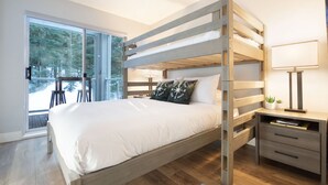 A modern bedroom with a wooden bunk bed, a bedside table with a lamp, and a view of the outdoors through a large window. - Mountainside 2Br/2Ba - Hot Tub/Pool/Free Parking (Whistler)