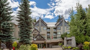 A multi-story hotel with a stone facade, multiple balconies, and a landscaped entrance area with trees and shrubs. - Mountainside 2Br/2Ba - Hot Tub/Pool/Free Parking (Whistler)