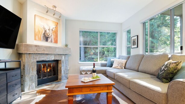 A living room with a stone fireplace, a large window, a sofa, and a television. - Mountainside 2Br/2Ba - Hot Tub/Pool/Free Parking (Whistler)