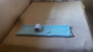 Un lit simple avec une tête de lit en bois, une couverture bleue et une serviette enroulée posée sur le lit.