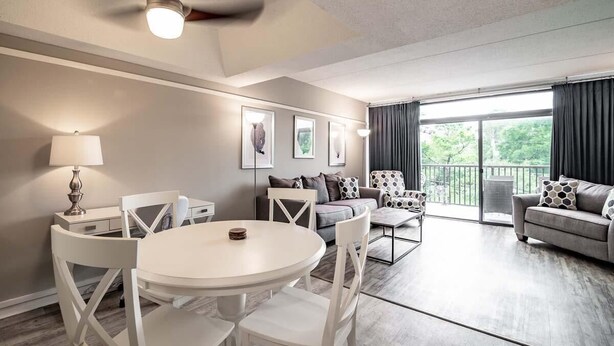 A modern living room with a white dining table, white chairs, a grey sofa, and a balcony with curtains. - Litchfield Retreat near Brookgreen Gardens (Pawleys Island)