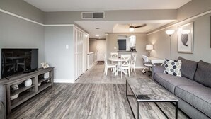 A modern living room with a grey sofa, a coffee table, and a TV on a wooden stand. There is a dining area with white chairs and a table, and a kitchen in the background. - Litchfield Retreat near Brookgreen Gardens (Pawleys Island)