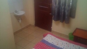 A bedroom with a bed, a small table, a sink, and a door. - Desai Annex Guest House (Nairobi)