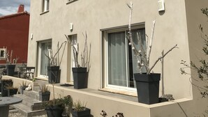 Een gebouw met twee verdiepingen, een beige gevel en een dak met rode tegels. Er zijn meerdere ramen, waarvan er sommige voorzien zijn van luiken, en verschillende plantenbakken met planten langs de buitenmuur.