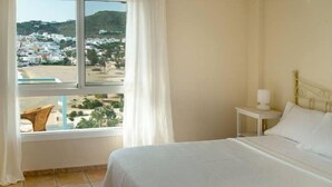 A bedroom with a bed, a nightstand, a lamp, and a view of a coastal town through a window. - Apartamento del Cabo I (Ventorros de San José)