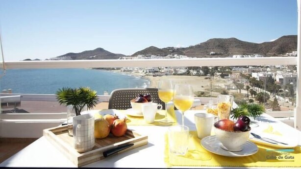A breakfast table with fruit, a cup, and a glass of orange juice, overlooking a beach and cityscape. - Apartamento del Cabo I (Ventorros de San José)